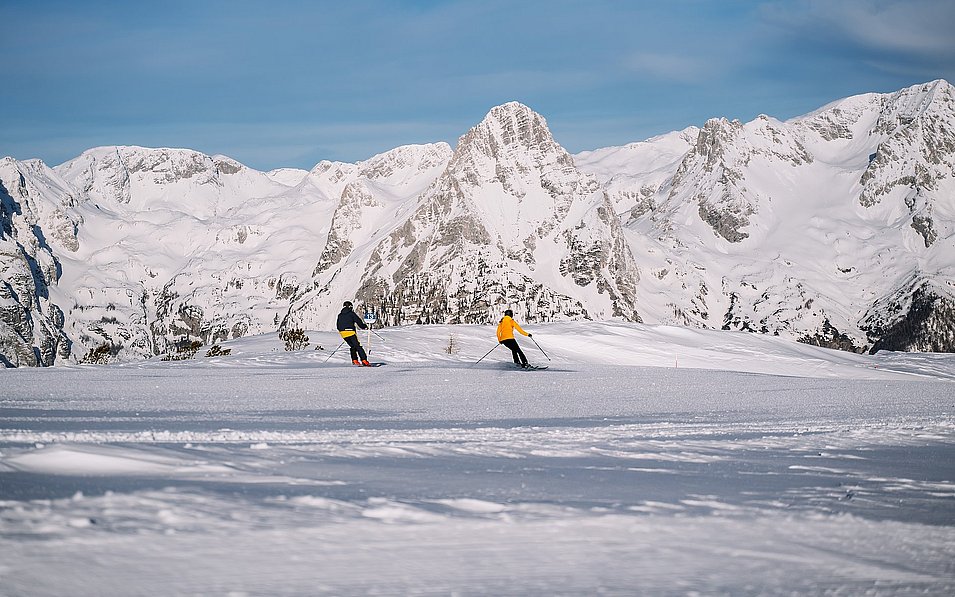 Ein Paar fährt die Piste auf der Höss in Hinterstoder hinunter. Im Hintergund erstreckt sich ein traumhaftes Bergpanorama.