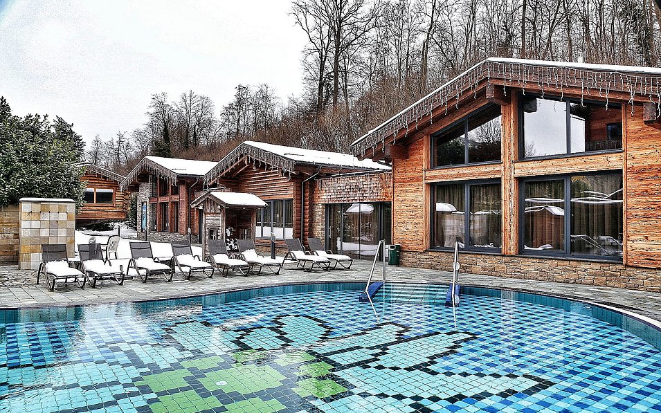 AusZeit – Das Sauna-Bergdorf. Blick auf die Außenanlage bei Tag im Winter. Die Liegestühle sind schneebedeckt.