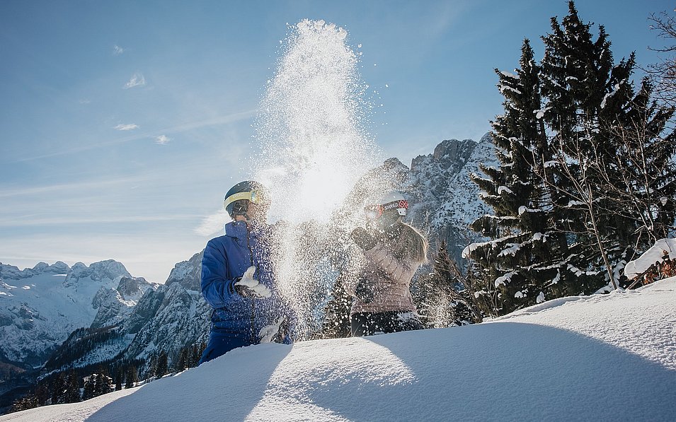 Eine Frau und ein Mann genießen den Winter. Sie werfen den frischen Pulverschnee in die Luft und die Sonne glitzert durch die Schneekristalle.