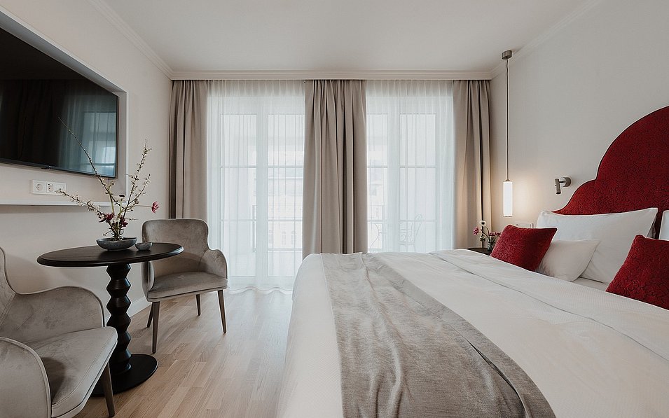 Blick in ein Classic Doppelzimmer mit Balkon im Hotel Grand Elisabeth in Bad Ischl im Salzkammergut, vorne das Bett und das TV Gerät an der Wand, im Hintergrund die Balkontür.