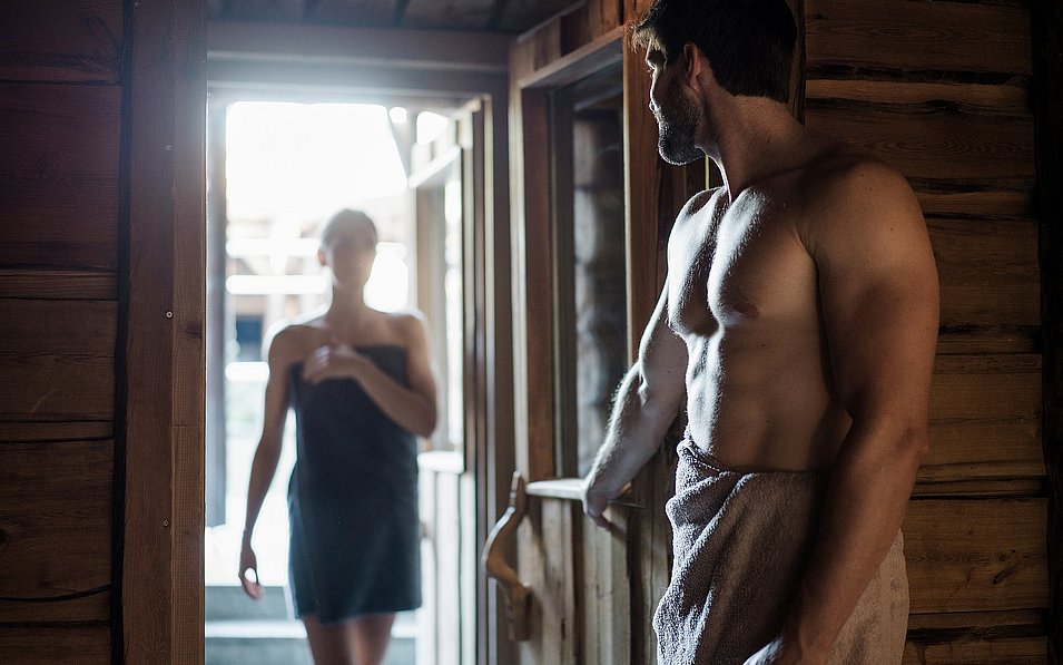 Ein Mann hält die Tür zu einer Sauna im Sauna Bergdorf AusZeit im EurothermenResort Bad Schallerbach auf, während von hinten eine Frau hereinkommt.